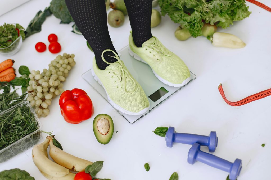 Person standing on scales surrounded by fresh produce and fitness equipment symbolizing a balanced lifestyle.